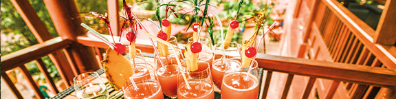 There are eight cocktails on a tray with pineapple, cherry and decorative sticks against a green background.