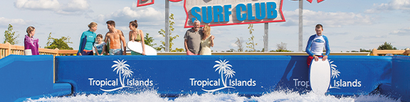 People with surfboards stand on a platform above a wave pool. Above them is a sign that says “Surf Club”.