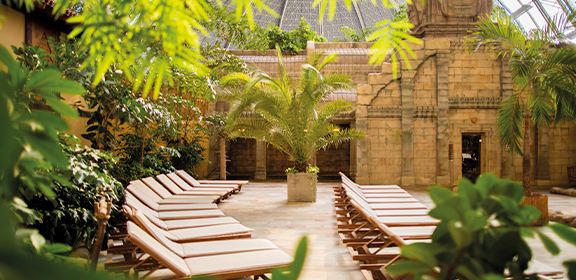 Tropically designed sauna area with loungers among palm trees and a view of a temple-like stone building.