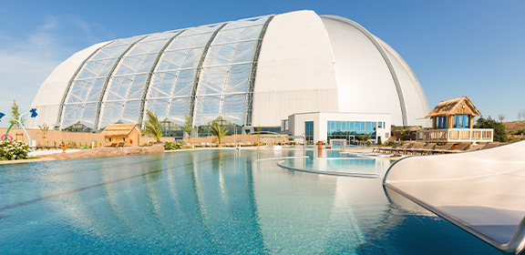 View of the outdoor pool at AMAZONIA with the striking white dome of Tropical Islands in the background under a blue sky.