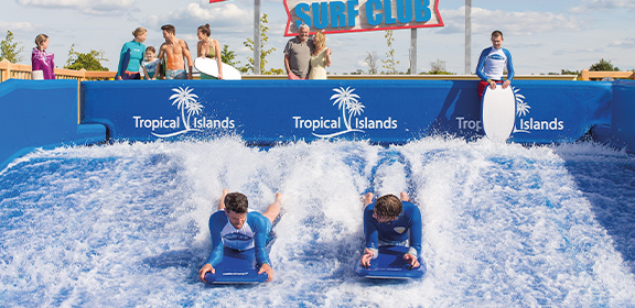 Two people bodysurfing in the Pororoca surf simulator while other people watch in the background.
