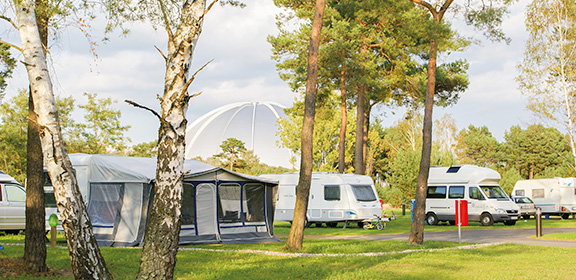 Campingplatz mit Wohnwagen, Zelten und weißer Kuppel im Wald bei Sonnenschein.