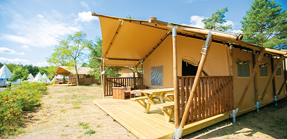 Large lodge tent with wooden terrace and picnic table on a natural campsite under a blue sky.
