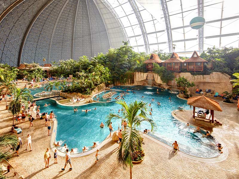 Tropical indoor pool surrounded by lush palm trees and exotic vegetation, with turquoise water, a whirlpool area, and relaxing lounge spaces beneath the iconic Tropical Islands dome