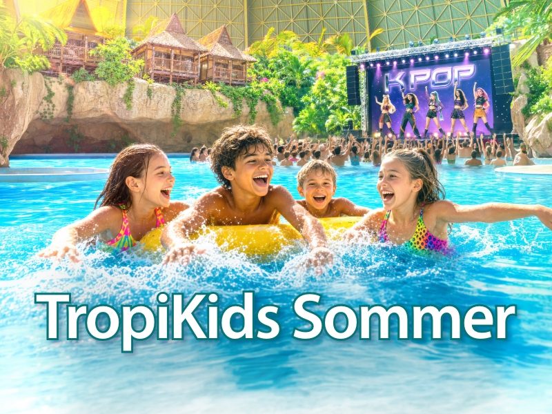 Four children play in a sunny outdoor pool, holding onto a large yellow inflatable ring in turquoise water; other swimmers and the pool edge are visible in the background.