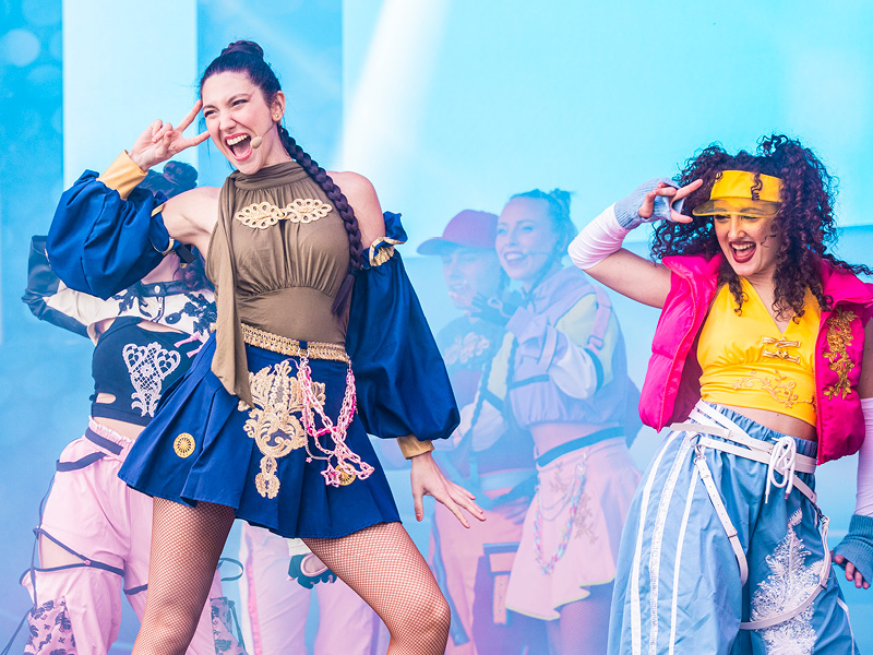 Dance group in colorful stage outfits performing an energetic choreography on a stage with a blue-lit background.