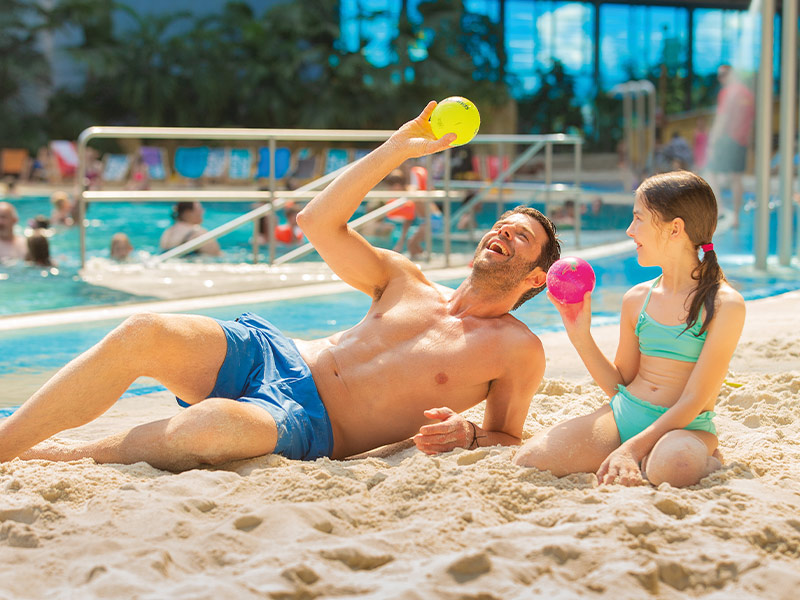 A man with a ball in his hand and a girl in swimwear sit on the sand with a pool of water in the background.