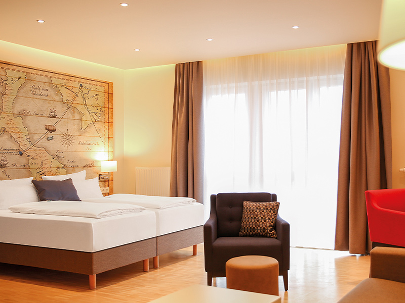 Bright comfort room with large bed, red chair, dark armchair, small stool and illuminated world map on the wall.
