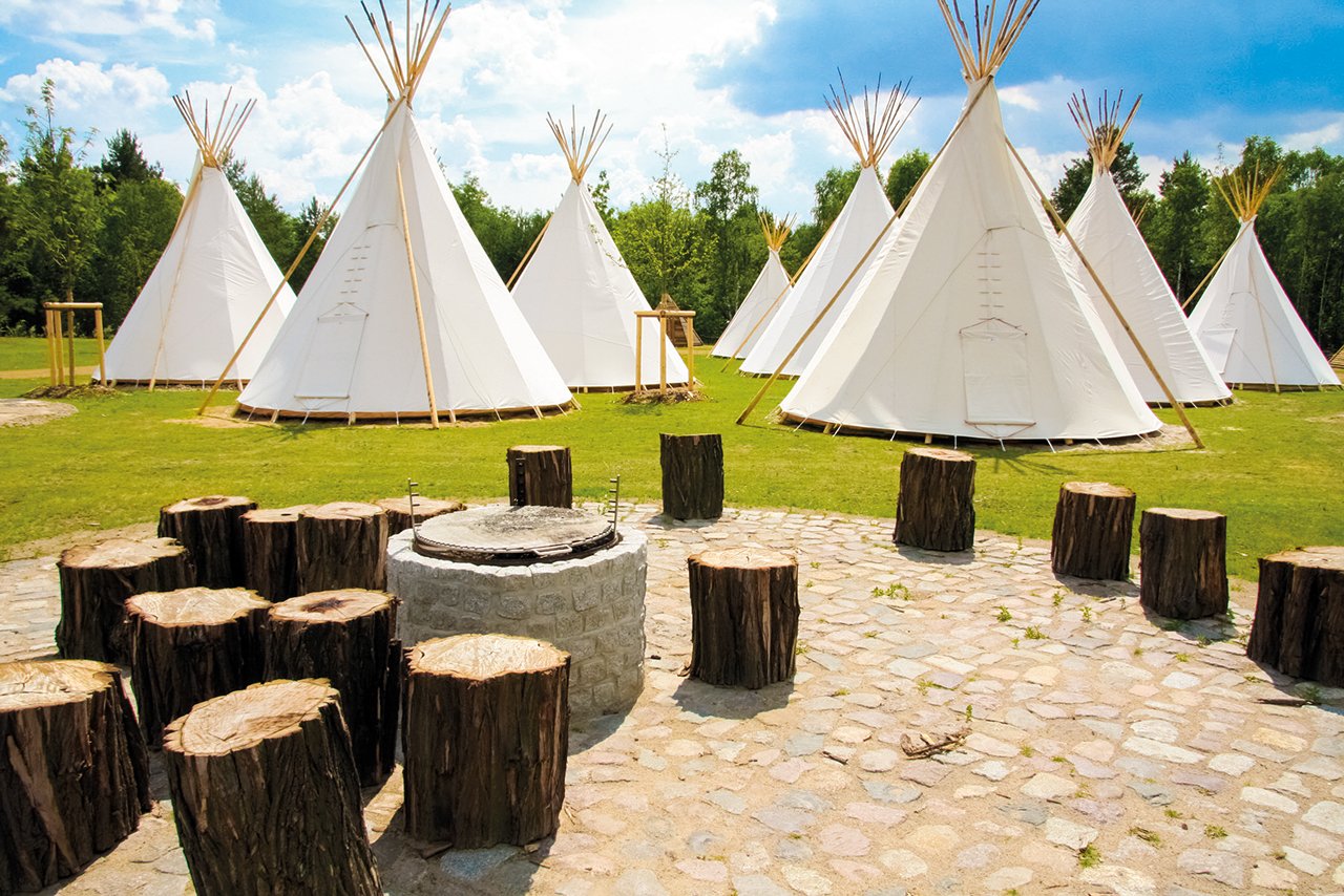 White tepees on a meadow under a partly cloudy sky, surrounded by trees.