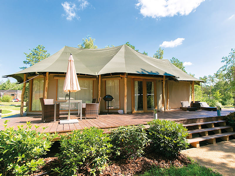 Large luxury tent with wooden terrace, garden furniture and surrounding greenery under a slightly cloudy sky.