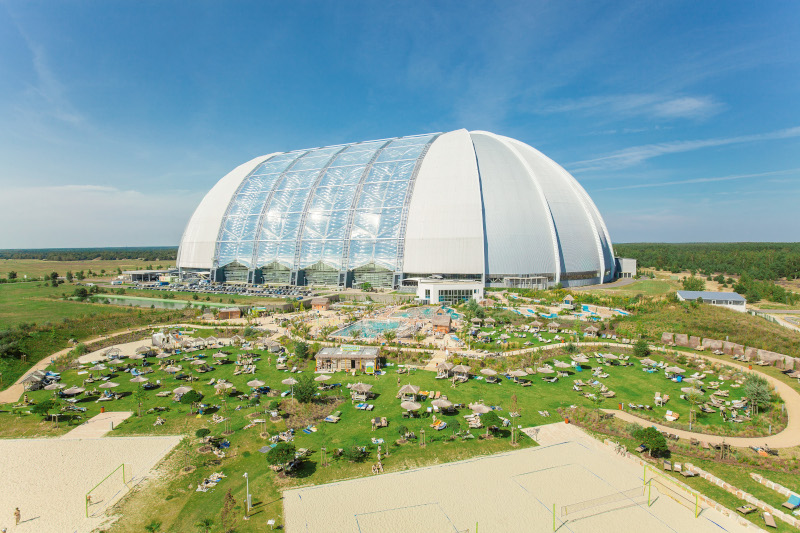 Sunbathing lawn with sun loungers and umbrellas in front of the large dome of the Tropical Islands hall.