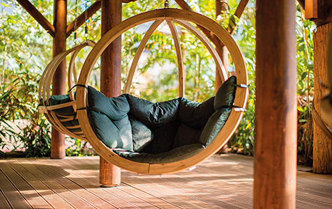 Wooden canopy with two suspended, upholstered round seats in a green outdoor setting.
