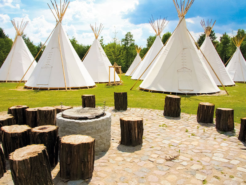 White tepees on a meadow under a partly cloudy sky, surrounded by trees.