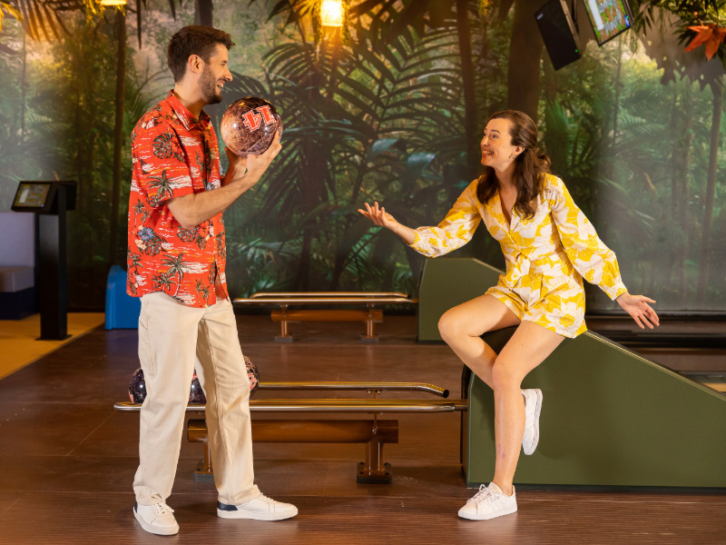 Two people in a bowling alley, one with a bowling ball, tropical decoration in the background.