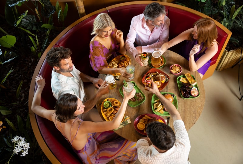 Six people at a round table with food and drinks, surrounded by greenery
