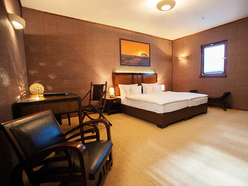A hotel room with brown walls and a large bed; in the foreground, dark chairs and a table.