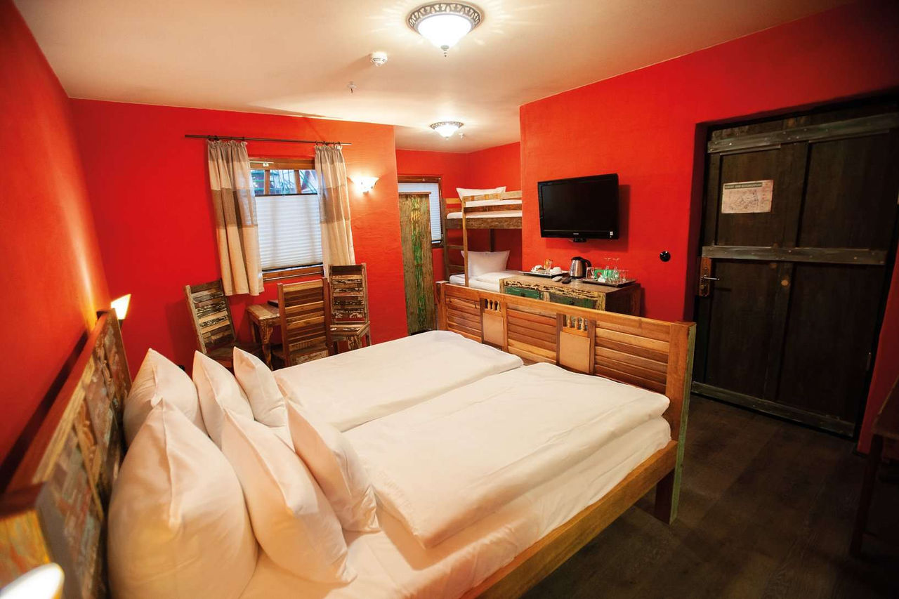 Cozy hotel room with red walls, double bed, bunk bed, TV, wooden furniture and windows with curtains.