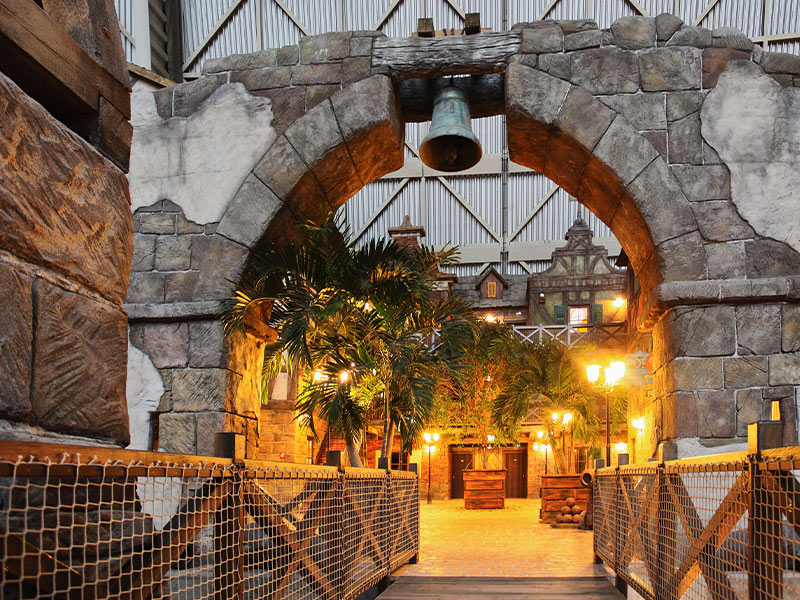 Stone arch with bell, leads into a tropically designed interior with wooden railings, decorative facades and street lamps.
